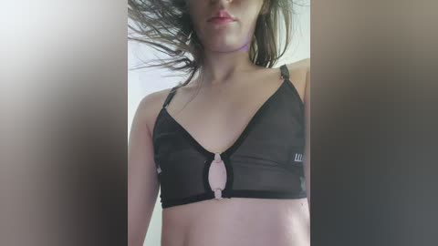 Media: Video of a light-skinned woman with dark hair, wearing a sheer black sports bra with a metallic ring detail, and a choker, against a plain beige background.