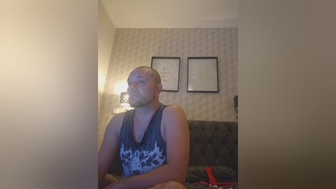 Video of a bald, muscular, light-skinned man in a grey tank top with a black pattern, sitting on a dark couch with a beige, patterned wallpaper behind him.