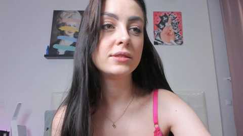 Video of a young woman with long, straight black hair, fair skin, and a pink bra, standing indoors with abstract art on the white wall behind her.