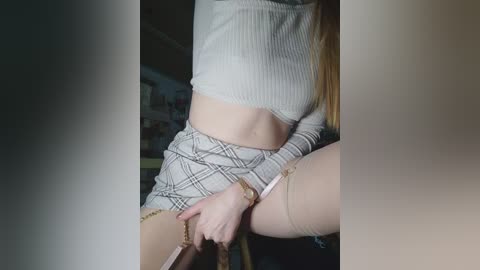 Video of a slim, fair-skinned woman with long blonde hair, wearing a ribbed crop top and plaid skirt, sitting on a chair, revealing her midriff and thighs.