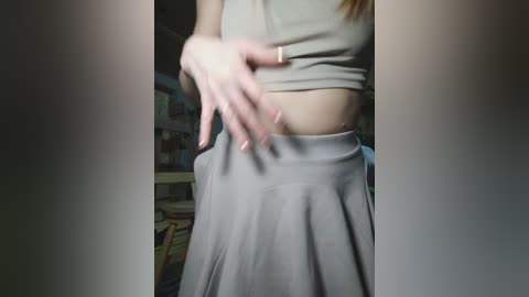 A video of a woman's torso in a light gray crop top and matching skirt, with a hand resting on her stomach, in a dimly lit room with bookshelves in the background.