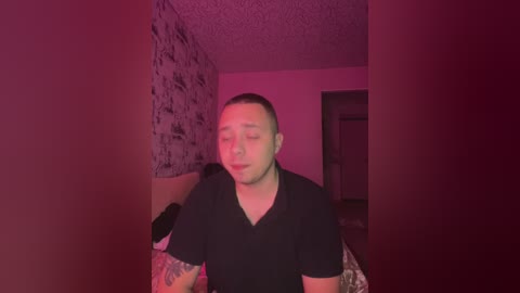 Video of a bald, light-skinned man with tattoos, wearing a black V-neck shirt, standing indoors with a red-hued background and patterned wallpaper.