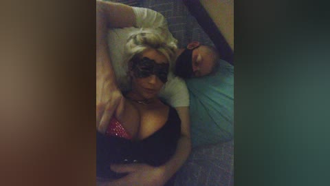 Video of a blonde woman in a black lace mask, lying on a bed next to a bald man, both sleeping, with a blurred background.