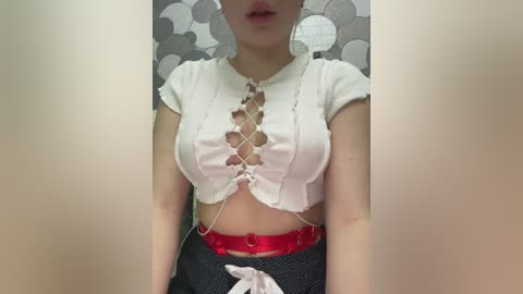 Media: Video of a young woman with fair skin, wearing a cropped white crop top with lacing and a red belt, against a gray and white patterned background.