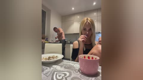 Media: Video of a blonde woman in a black top, eating strawberries at a kitchen table. Two shirtless men are cooking in the background.