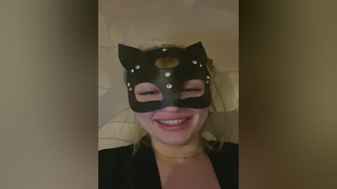Video of a young woman with light skin, smiling, wearing a black cat mask with white studs, and a black top, against a beige wall.