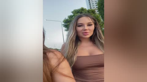 Media: Video of a young, fair-skinned woman with long, wavy blonde hair, wearing a strapless brown dress, standing outdoors near a green tree and a high-rise building.