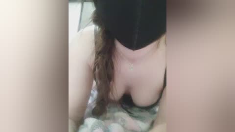 A close-up video of a topless woman with long brown hair, wearing a black headband, lying on a patterned blanket. The image is slightly blurred and grainy, focusing on her chest and neck.