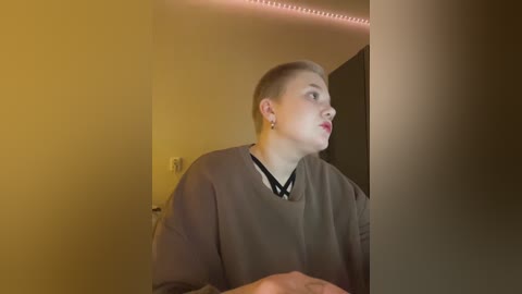 A video of a person with short, light blonde hair, fair skin, and a round face, wearing a grey sweater, looking upwards in a dimly lit room with yellowish lighting and a red LED strip along the ceiling.