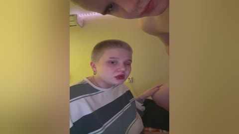 Video of a young woman with short, platinum blonde hair, wearing a striped t-shirt and red lipstick, standing in a dimly lit room with yellow walls, capturing a reflective mirror image of her face.