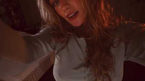 Media: Video of a smiling, light-skinned woman with wavy, long auburn hair, wearing a button-up shirt. She extends her arm, capturing the moment.