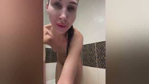 A video of a young, fair-skinned woman with wet, dark hair, standing in a modern bathroom with beige and black mosaic tiles. She is topless, smiling slightly, and has a nose ring.