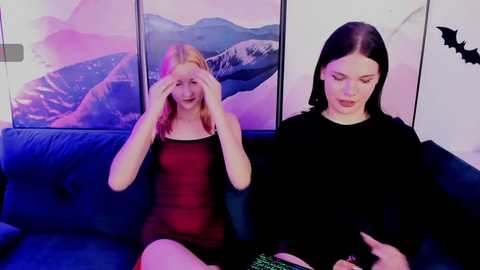 Video of two young women sitting on a dark blue sofa in a room with bat wall art; one wears a sheer red dress, the other a black sweater, both with pale skin.