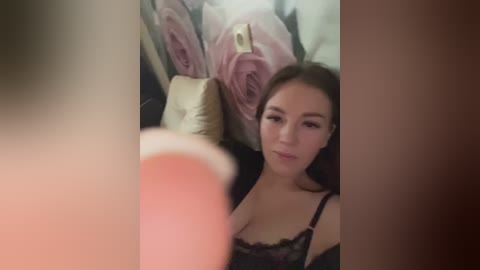 Video of a young Asian woman with long brown hair, wearing a black lace bra, sitting on a bed with floral pillows, blurred foreground.
