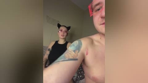 Video of a fair-skinned, topless woman with a tattoo sleeve and cat ears, standing beside a shirtless man with a red bandana. Both appear indoors with beige walls.