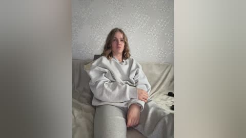 Video of a Caucasian woman with wavy, shoulder-length blonde hair, wearing a gray sweatshirt and matching sweatpants, sitting on a beige bed with floral-patterned wallpaper in the background.