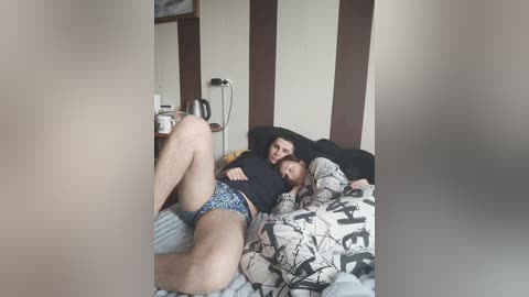 A video of a man and woman cuddling on a bed, wearing patterned boxer shorts. The room has a cluttered desk with a lamp and books.