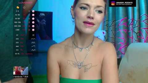 A video of a young woman with fair skin and dark hair, wearing a green strapless dress, silver necklace, and butterfly tattoo on her chest. Background includes a blue wall, pink curtains, and a black screen.