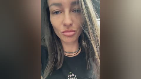 Media: Video of a young woman with light skin, straight brown hair, and light makeup, wearing a black t-shirt and multiple choker necklaces. She has a neutral expression and is indoors.