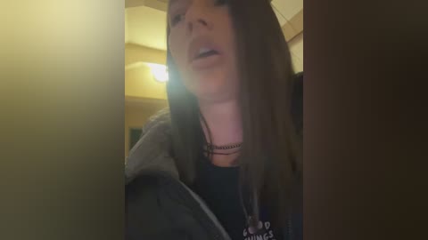 Media: Video of a young woman with long brown hair, wearing a black hoodie and necklace, standing in a dimly lit room with yellow walls and a lamp.