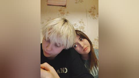 Media: Video of two young people with light hair, one with platinum blonde, the other with brown. They're sitting close, both looking at the camera. The background features floral wallpaper and a pink towel.