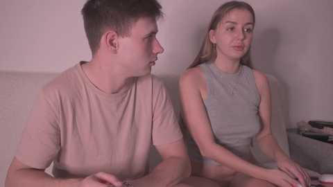 A video of a young Caucasian couple sitting side-by-side on a beige couch. The man, with short brown hair, wears a plain beige T-shirt. The woman, with long blonde hair, wears a gray tank top. They have a neutral expression.