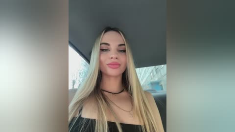 Media: Video of a young, light-skinned woman with long, straight blonde hair and fair skin, wearing a black off-shoulder top and a black choker necklace, standing indoors with a blurred background of a gray room.