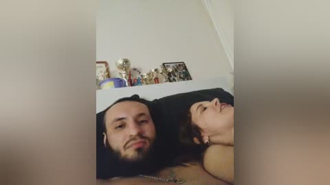 Video of a bearded man and a woman with short hair, lying together on a bed, smiling. Background features a white wall with framed pictures and a blue vase.