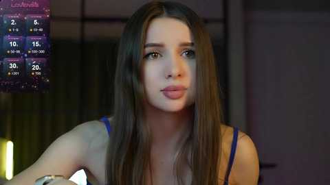 Media: Video of a young Caucasian woman with long brown hair, wearing a blue tank top, sitting indoors with dim lighting, displaying a digital calendar on the left.