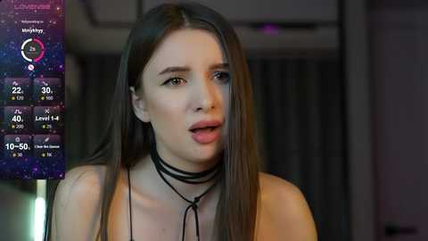 Media: Video of a young woman with long brown hair, wearing a black choker and top, against a dimly-lit room background.