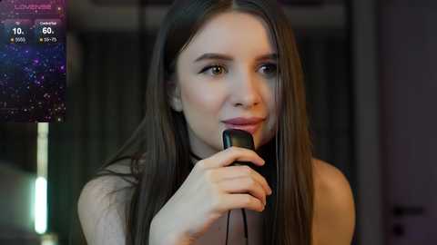 Media: Video of a young woman with long brown hair, wearing a white top, holding a black cellphone to her ear. Background features a dimly lit, blurred room.