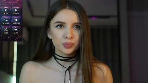 Media: Video of a young woman with long, straight brown hair, fair skin, and brown eyes, wearing a black choker necklace. Background shows a dimly lit room with a calendar display.