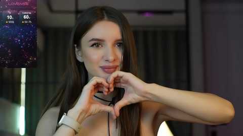 Media: Video of a young woman with long, straight dark hair, fair skin, and a slim physique, forming a heart shape with her hands in front of her face, smiling. She wears a white top and a black choker necklace. Background shows dimly lit, modern interior.