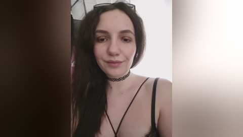A video of a young woman with pale skin, dark hair, and a black choker necklace, wearing a black lace bra. She has a slight smile, and the background is blurred.