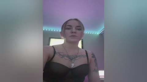 Media: Video of a fair-skinned woman with blonde hair, wearing a black sheer top revealing tattoos on her chest, standing in a dimly lit room with purple and blue LED lighting.