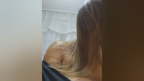 Media: Video of a woman with long, straight blonde hair, partially covered by a black garment, sitting in a dimly lit room with white walls and a white curtain.