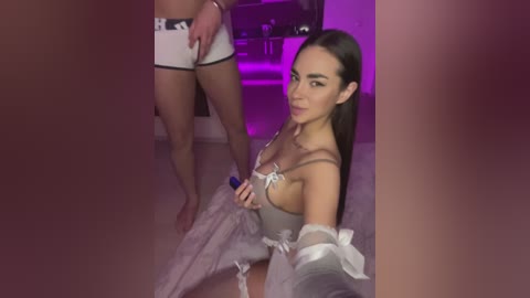 Media: Video of a young woman with long brown hair and fair skin, wearing a sheer, lavender dress, kneeling on a wooden floor. A man, wearing white boxer shorts and a black belt, stands in the background. The room is dimly lit with purple lighting.