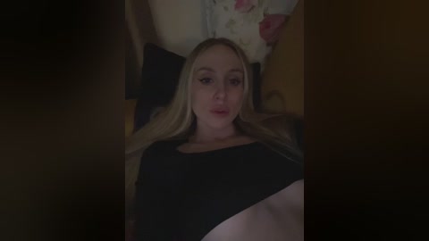 Video of a young, fair-skinned, blonde woman with long hair, wearing a black top, lying on a bed with dim lighting, floral pillowcase, and dark sheets.