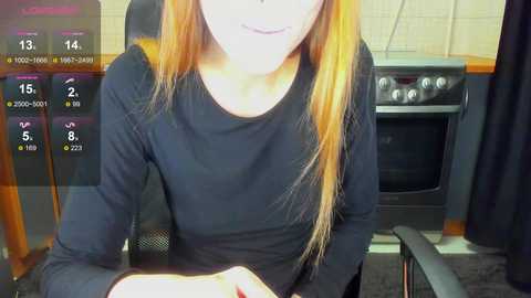 Video of a young woman with long orange hair, wearing a black long-sleeve shirt, seated at a table in a kitchen with a black oven and white tiled walls, overlaid with virtual game interface.