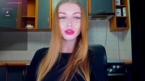Video of a blonde woman with long hair, wearing black lipstick, sitting in a modern kitchen with wooden cabinets and white tiled backsplash.