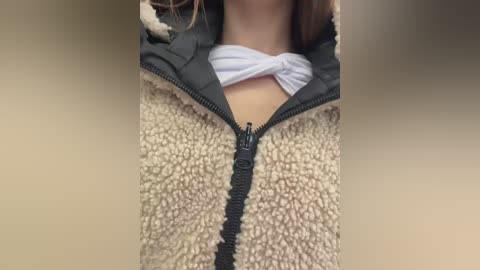 Media: Video of a woman's torso wearing a beige, textured fleece jacket with a black zipper over a white shirt tied at the neck, partially visible.