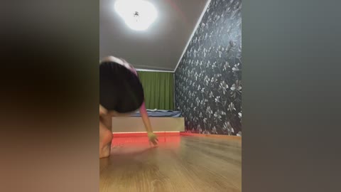 Media: Video of a person with a blurred face, wearing a black shirt and pink shorts, performing a handstand on a wooden floor. The room features a green curtain, a black patterned wall, and a bright ceiling light.