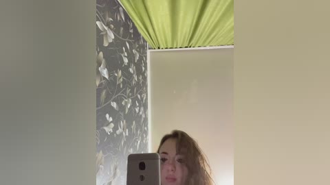 Media: Video of a young woman with light skin and shoulder-length blonde hair, partially visible, taking a selfie in a room with a green ceiling, floral wallpaper, and beige walls.