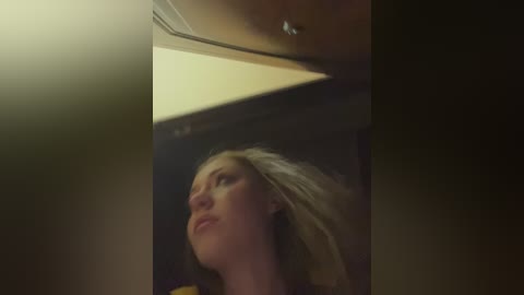 Media: Video of a blonde woman with fair skin, wearing a yellow top, looking upward. The background is dimly lit, with a ceiling light above her. Her hair is tousled, and she appears contemplative.