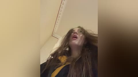 Media: A video of a young woman with long, messy brown hair, wearing a black jacket with yellow accents, standing in a dimly lit room with beige walls and a white ceiling.