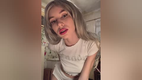 Video of a young woman with long, platinum blonde hair, wearing a white crop top and matching panties, sitting in a cozy, softly lit room with floral curtains and a wooden chair in the background.