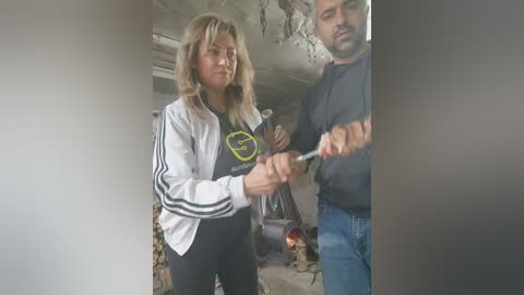 Media: Video of a blonde woman in a white jacket with black stripes and black leggings, holding a hammer against a man in a hoodie and jeans, in a dimly lit room with a fireplace and wooden beams.