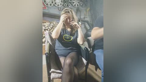 Media: Video of a blonde woman in a dark shirt, shorts, and black tights, sitting on a wicker chair, covering her face with her hands, surrounded by wooden objects in a rustic room.