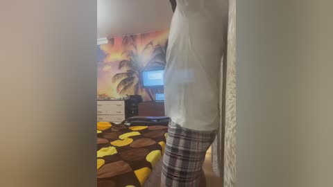 Video of a person standing in a bedroom with a white tank top and plaid pajama pants. Background features a tropical-themed mural, a dresser, and a bed with a brown comforter adorned with yellow and orange shapes.