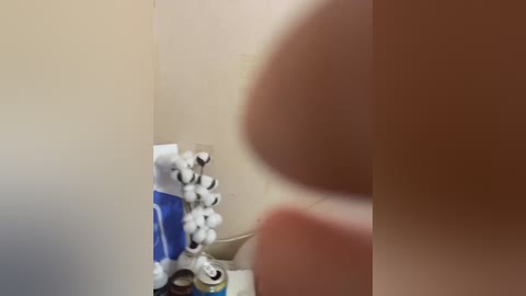 A close-up video of a woman's hand holding a white tube of toothpaste with a blue lid, positioned against a beige wall. The image is slightly blurred, emphasizing the hand and tube.